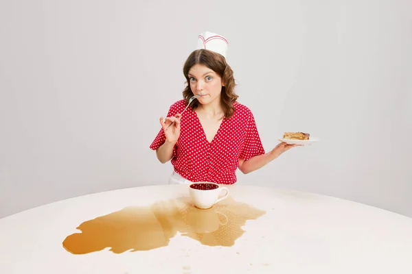 Stylish young female waitress in retro american fashion style of 70s, 80s sitting at table and drinking coffee over grey background. Funny meme emotions, ad, sales. Weird people concept