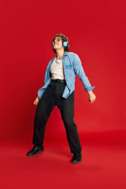 Happiness mode. Portrait of young excited man, student in headphones dancing isolated on red background. Concept of positive emotions, youth culture, freedom, active lisfestyle.