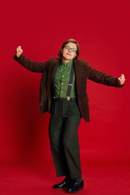 Social dance. Studio footage young man, student or office clerk in vintage fashion style costume, suit posing isolated on dark red background. Positive emotions, delight, art, style