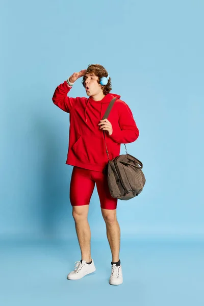 Young sportive man in sportswear holding huge sportive bag isolated over blue background. Concept of active healthy lifestyle, positive emotions, sport, fitness, ad. Model looks happy, delighted