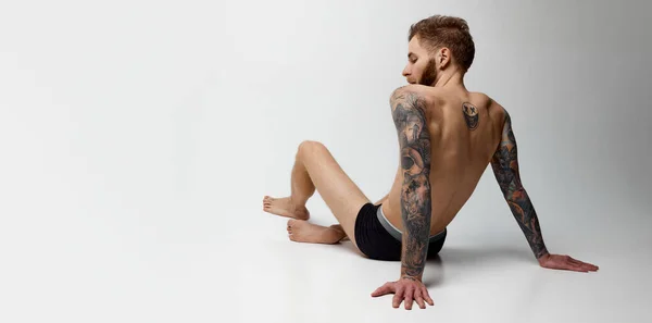 Studio shot of young blonde man with beard and moustache posing shirtless. Muscular body shape. Tattoo body art. Concept of men fashion, style, body aesthetics, beauty, mens health and emotions