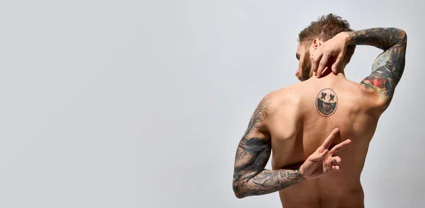 Studio shot of young blonde man with beard and moustache posing shirtless. Muscular body shape. Tattoo body art. Concept of men fashion, style, body aesthetics, beauty, mens health and emotions