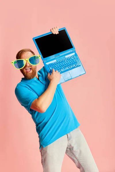 Cheerful funny Caucasian man wearing casual style clothes having fun, working with laptop over pink background. Positive emotions, education, happiness. Complementary colors