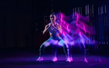 Squats. Sportive woman in sportswear and sportsfoot training in modern fitness room over dark background with mixed neon light. Active and healthy lifestyle concept
