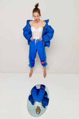 Portrait of beautiful woman in retro sports style trousers and down jacket posing with mirror over light background. Concept of weird people, fashion, creativity, aspiration. Copy space for ad