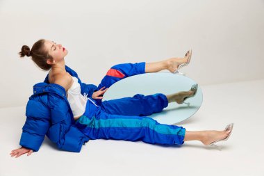 Portrait of beautiful woman in retro sports style trousers and down jacket posing with mirror over light background. Concept of weird people, fashion, creativity, aspiration. Copy space for ad