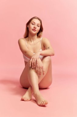 Relax time. Young sensual woman wearing white inner wear sitting on floor over pink color background. Concept of natural beauty, body and skin care, healthy lifestyle. Copy space for ad