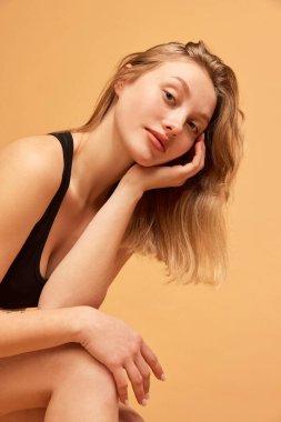 Charming girl with long blonde hair. Portrait of young slim beautiful girl wearing black underwear posing over sand color background. Concept of beauty, body and skin care, healthy eating