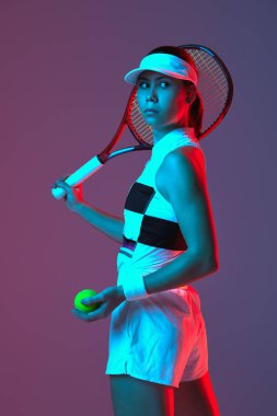 Young sportive girl professional tennis player posing with tennis racket gradient pink-purple background in neon. Concept of sport, strength, professional skills, active lifestyle and beauty, ad.
