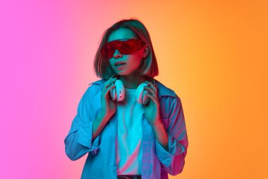 Merak ve ilgi. Genç duygusal kız, neon ışıkta pembe-turuncu arka planda poz veren şık bir göz giysisi giyiyor. Duygu, gençlik, moda, ruh hali ve müzik kavramı.