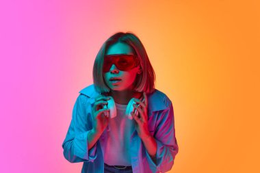 Merak ve ilgi. Genç duygusal kız, neon ışıkta pembe-turuncu arka planda poz veren şık bir göz giysisi giyiyor. Duygu, gençlik, moda, ruh hali ve müzik kavramı.