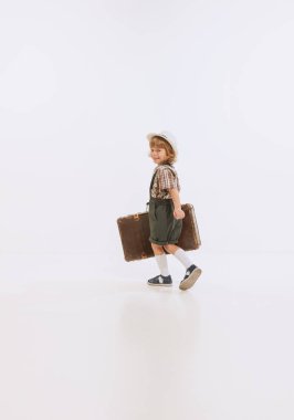 Smiling little boy, kid wearing retro style clothes with huge retro suitcase isolated over white background. Concept of emotions, 60s vintage fashion style, childhood, leisure activities, vacation