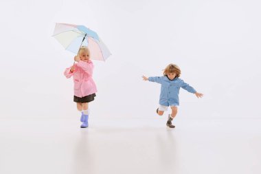 Cute children, happy kids in raincoats playing, having fun together isolated over white background. Boy and girl with unmbrella. Concept of childhood, friendship, family, fun, joy, fashion