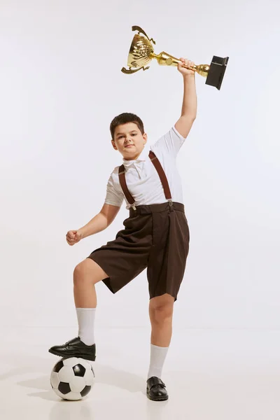 Win. Studio shot of little boy, preschool age kid wearing retro style clothes holding winners cup isolated over white background. Concept of emotions, childhood, hobby, success, achievement