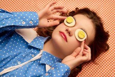 Strange look. Weird people concept. Young girl lying on table with eggs on her eyes over peach color background. Vintage, retro style. Food pop art photography, fashion, kitsch concept