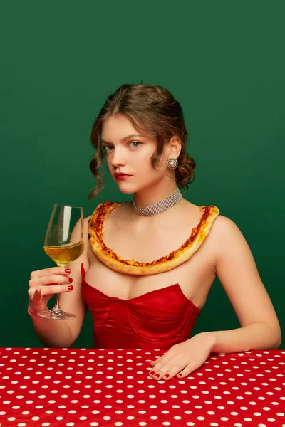 Tasting wine. Young stylish beautiful girl with pizza crust on her shoulders sitting at table over green background. Vintage, retro style. Complementary colors. Food pop art photography.