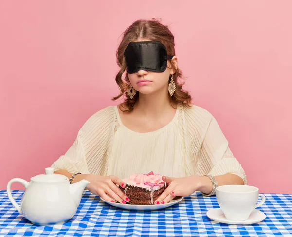 Young girl with tousled hair after sleep and with sleep mask eating cake, pie and drinking tea. Concept of emotions, weird people, retro fashion and humor. Food pop art photography
