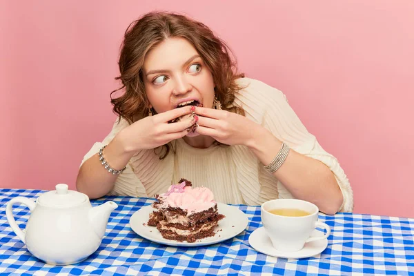 Emotional young girl with tousled hair after sleep greedily eating cake, pie and drinking tea over pink background. Concept of emotions, weird people, retro fashion and humor. Food pop art photography