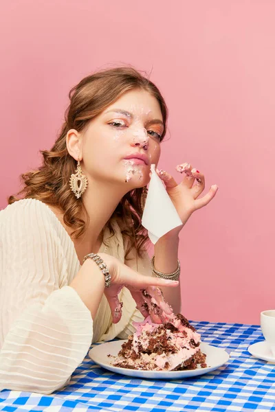 Out of etiquette. Charming young girl eating cake, pie and drinking tea. Sweet cream mask. Concept of emotions, weird people, retro fashion and humor. Food pop art photography