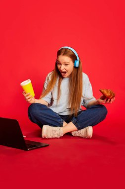 Surprised young girl, student wireless headphones sitting on floor, studying isolated over magenta color background. Concept of education, studying, homework, youth, lifestyle