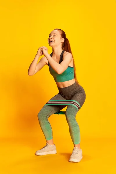 Squats. Beautiful girl in sports fitness clothes doing exercises with equipment over yellow background. Sport, fitness, happy positive emotions and active lifestyle concept. Copy space for ad
