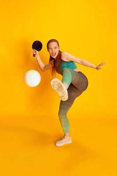 Cute funny young girl in sports fitness clothes playing ping-pong tennis over yellow background. Sport, fitness, happy positive emotions and active lifestyle concept. Copy space for ad