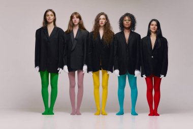 Diversity. Group of young diverse women in black jackets and multi colored tights standing together over grey background. Concept of beauty, feminism, emotions, extraordinary fashion, modern style