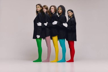 Diversity. Group of young diverse women in black jackets and multi colored tights standing together over grey background. Concept of beauty, feminism, emotions, extraordinary fashion, modern style