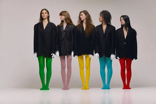 Art, beauty. Studio shot of young stylish beautiful fashion models in black jackets and multi colored tights posing over grey background. Concept of feminism, extraordinary fashion, modern style