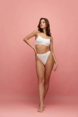 Young tall thin girl with long soft brown hair wearing white inner wear posing over pink color background. Concept of natural beauty, body and skin care, healthy eating, anti-cellulite program, spa.