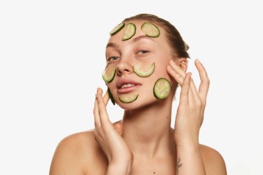 Revival facial mask. Adorable young woman with perfect skin and slices of fresh cucumber on her face isolated on white background. Beauty, skin care, cosmetology, spa, ad concept