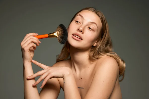 Morning beauty procedure. Young sensual woman with bare shoulders powdering with makeup brush over grey background. Concept of beauty, natural make-up, skin care, healthy look