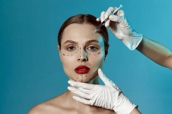 Young beautiful woman. Plastic surgery or face lifting for young female. Doctors hands wearing gloves drawing lines, holding head of patient, isolated on blue. Rhinoplasty, rejuvenation concepts