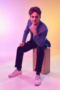 Portrait of young handsome young man, office clerk, student in jacket and jeans posing over light gradient yellow-purple background. Fashion, art, education, business concept. Human emotions