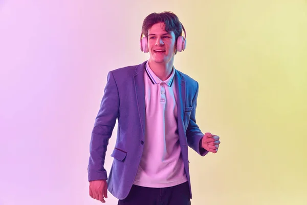 Music. Half-length portrait of young man in headphones dancing isolated on gradient yellow-purple background in neon light. Concept of human emotions, youth culture. Copy space for ad.