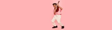 Happy emotional little african boy in retro style clothes and cap posing with ukulele guitar isolated on pink background. Concept of music, childhood, education, fashion and aspiration. Banner