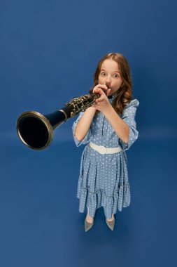 Monochrome portrait of charming nice little girl in dress and big shoes playing on clarinet over blue background. Music, fashion, hobby concept. Child with retro musical instrument. Wide angle view
