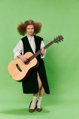 Beautiful little girl, funny musician wearing huge oversize clothes playing guitar, having fun isolated over green background. Pop art, music, new vision, fun concept. Aspiration and creativity