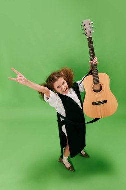 Beautiful little girl, funny musician wearing huge oversize clothes playing guitar, having fun isolated over green background. Pop art, music, new vision, fun concept. Aspiration and creativity