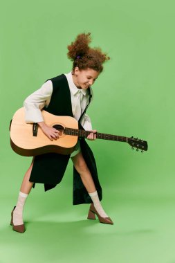 Beautiful little girl, funny musician wearing huge oversize clothes playing guitar, having fun isolated over green background. Pop art, music, new vision, fun concept. Aspiration and creativity