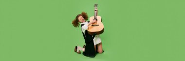 Beautiful little girl, funny musician wearing huge oversize clothes playing guitar, having fun isolated over green background. Pop art, music, new vision, fun concept. Banner, flyer for ad