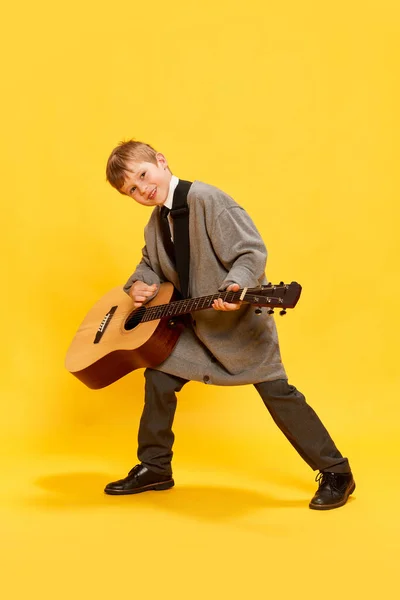 Charming little boy, funny musician wearing huge oversize clothes playing guitar, having fun isolated over yellow background. Pop art, music, new vision, fun concept. Aspiration and creativity
