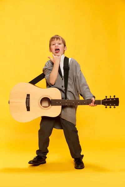Charming little boy, funny musician wearing huge oversize clothes playing guitar, having fun isolated over yellow background. Pop art, music, new vision, fun concept. Aspiration and creativity