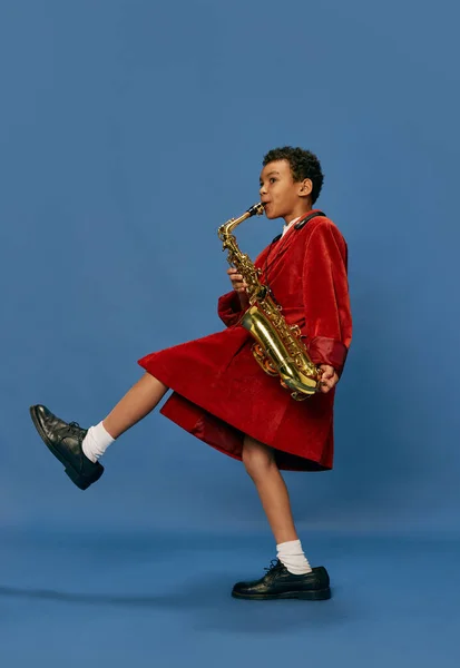 Like jazz man. Portrait of cute little african boy wearing huge mans jacket and shoes playing on saxophone over blue background. Fashion, art, music and creative style. Looks happy, cheerful