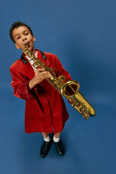 Like jazz man. Wide angle view image of cute little african boy wearing huge mans jacket and shoes playing on saxophone over blue background. Fashion, art, music and creative style. Melody