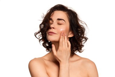 Feeling of freshness. Young sensual woman with bare shoulders apply face cream over white background. Fashion, beauty, ad, spa, cosmetology, skincare concept.