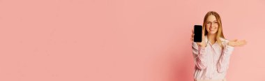 Happy young pretty blonde girl, student wearing modern office style clothes showing phone screen over pink background. Concept of youth, student college life, business and education. Banner for ad