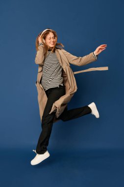 Adorable caucasian young woman in wireless headphones listening to music and dancing on blue navy background. Blonde girl in coat smiles. Concept of happiness, fashion, youth, ad and music