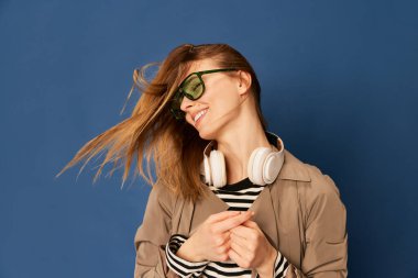 Adorable caucasian young woman in wireless headphones listening to music and dancing on blue navy background. Blonde girl in coat smiles. Concept of happiness, fashion, youth, ad and music