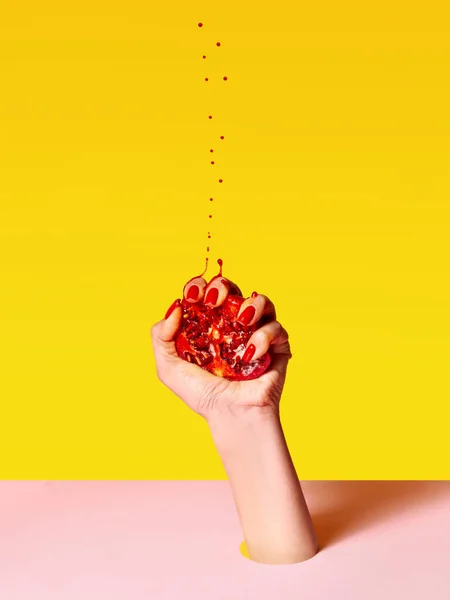 Making juice. Human hand with red painted nails squeezing half of pomegranate over pink yellow background. Contemporary art. Drops of juice fly up. Concept of healthy eating, fashion, ad